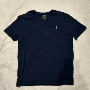 Mörkblå t-shirt Polo Ralph Lauren - Mörkblå t-shirt från Polo Ralph Lauren med v-ringning och klassisk broderad logga på bröstet. Tillverkad i mjuk tjock högkvalitativ bomull och har en normal passform. Perfekt för en clean och stilren look.
