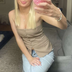 Zara rutig tubtopp med knappar - Snygg beige tubtopp med rutigt mönster och dekorativa knappar framtill. Toppen är figurnära och har en liten slits nertill framtill. Perfekt att styla med jeans för en trendig look.