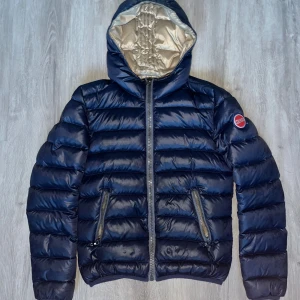 Mörkblå Colmar dunjacka med huva - Snygg mörkblå dunjacka från Colmar med glansig finish och klassisk logga på ärmen. Jackan har huva, två dragkedjeförsedda fickor och quiltad design. Insidan är beige och materialet känns lätt men ändå varmt. Perfekt för kyliga dagar. Pris kan diskuteras 