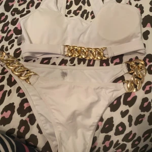 Vit bikini med guldkedjor, storlek L - Superfräsch vit bikini med breda guldfärgade kedjedetaljer både på överdelen och sidorna av underdelen. Sportig topp med breda axelband och högt skurna trosor. Materialet är nylon, polyester och spandex för en skön och stretchig känsla.