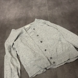 Ljusgrå kofta med knappar - En stilren ljusgrå kofta med långa ärmar och knappar framtill. Koftan har ribbade muddar vid ärmslut och nederkant, vilket ger en snygg och enkel look! Koftan är använd men i bra skick