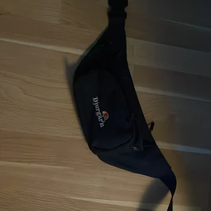 Svart Djurgårn axelväska med dragkedja - Säljer en svart axelväska från Djurgårn med justerbart band och praktisk dragkedja. Väskan har ett broderat Djurgårn-tryck med färgdetalj på framsidan och en smidig ficka för dina viktigaste grejer. Perfekt för dig som vill ha en sportig och enkel look.