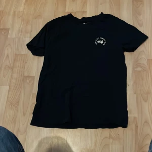 Svart t-shirt med tryck - Svart t-shirt med rund hals och korta ärmar. På bröstet finns ett litet vitt tryck med texten 'Chins up, shapeless no' och en illustration av en hantel. Materialet är mjuk bomull, perfekt för en chill och enkel stil.