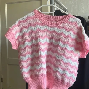 Rosa och vit stickad tröja med zigzag - Supergullig stickad tröja i rosa och vitt med vågigt zigzag-mönster. Tröjan har korta ärmar, rund halsringning och bred ribbad mudd nedtill. Perfekt för dig som vill ha en söt och unik look till våren eller sommaren!