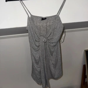 Grå kort klänning från Gina Tricot - Säljer en grå topp från Gina Tricot med smala axelband och snygg knytning och har en avslappnad passform. Perfekt för varma dagar eller när du vill ha något enkelt men stilrent.