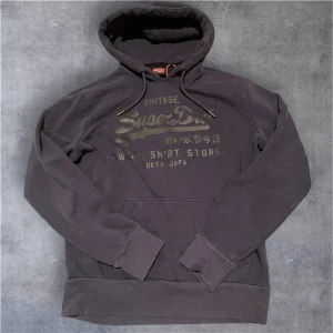 Mörkblå Superdry hoodie  - Säljer en mörkgrå hoodie från Superdry med stort tryck på bröstet och japanska tecken. Tröjan har huva med snörning, ribbade muddar och en klassisk känguruficka framtill. Perfekt för dig som gillar streetwear och vill ha en chill look.