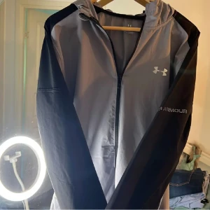 Grå och svart vindjacka Under Armour - Snygg vindjacka från Under Armour i grått och svart med huva och hel dragkedja framtill. Jackan har sportig look, logga på bröstet och är tillverkad i lätt polyester. Perfekt för aktiva dagar och streetstyle.
