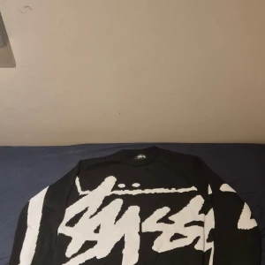 Svart och vit stickad Stüssy tröja - Säljer en svart stickad tröja från Stüssy med stort vitt grafiskt tryck över hela framsidan och baksidan. Tröjan har rund hals, ribbade muddar och lång ärm. Perfekt för dig som gillar streetwear och vill sticka ut med en fet logga.
