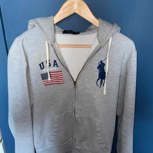 Grå hoodie från Polo Ralph Lauren - Säljer en grå hoodie från Polo Ralph Lauren med dragkedja och huva. Framsidan har USA-flagga, blå Polo-logga och text. 