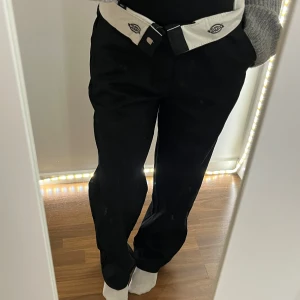 Svarta Dickies byxor med vit detalj - Svarta byxor från Dickies med bred passform och vit kontrastdetalj i midjan där loggan syns. Klassisk look med raka ben och skön loose fit, perfekta för en avslappnad streetstil.