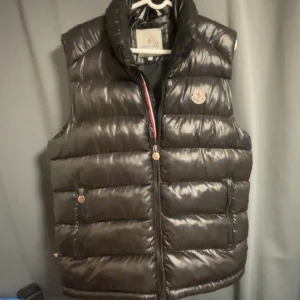 Svart dunväst från Moncler - Snygg svart dunväst från Moncler med glansig finish och klassisk logga på bröstet. Västen har hög krage, två fickor med dragkedja och quiltad design. Perfekt för lager-på-lager och ger en clean streetstyle-vibe.