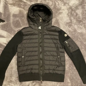 Svart Moncler cardigan - Svart pufferjacka från Moncler med stickade ärmar och quiltad kropp. Jackan har huva med snörning, dragkedja fram och fickor med tryckknappar. Moncler-logga på ärmen och extra ficka med dragkedja. Perfekt för dig som gillar stilren och sportig look.