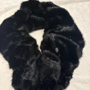 Svart fluffig halsduk från Laura Plus - Säljer en svart, supermjuk och fluffig halsduk från Laura Plus. Halsduken är gjord av akryl och polyester och har en pälsliknande yta som känns lyxig och varm. Perfekt att slänga på sig när det är kallt ute. Enkel att stänga med diskret krok.