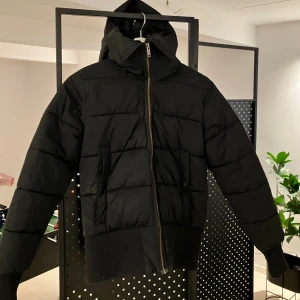 Svart puffer/dunjacka från Everest - Svart puffer/dunjacka från Everest med huva, ribbade muddar och två fickor med knappstängning. Jackan har en grov dragkedja framtill och quiltad design. Perfekt för kalla dagar och har en clean, modern look.