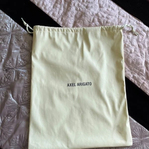 Gul dustbag från Axel Arigato - Säljer en ljusgul dustbag från Axel Arigato med dragsko upptill. , stilren design med svart logga framtill. Perfekt för att förvara sneakers eller accessoarer.