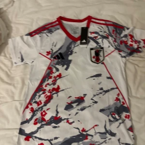 Japan landslagströja Adidas M - Japan landslagets officiella fotbollströja från Adidas i slim fit. Tröjan är vit med grå och röda abstrakta mönster, röda detaljer vid krage och axlar samt broderad landslagsemblem och Adidas-logga. Tillverkad i lätt polyester, perfekt för match eller träning.