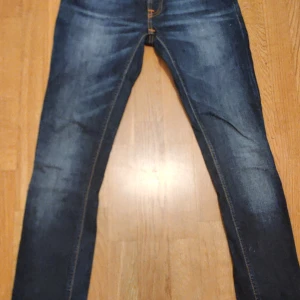 Mörkblå jeans från Nudie Jeans - Snygga mörkblå jeans från Nudie Jeans med klassiska orange sömmar och broderade bakfickor. Jeansen har en smal passform och är tillverkade i slitstarkt denim. Perfekta för dig som gillar en stilren och tidlös look.