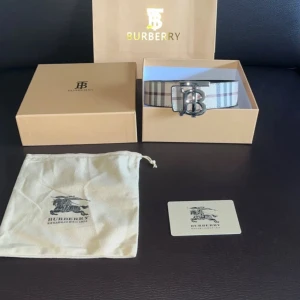 Burberry rutig bälte med silverdetalj - Snyggt Burberry-bälte med klassiskt rutigt mönster i beige, grått och vitt. Bältet har en stor silverfärgad spänne med Burberrys logotyp. Perfekt accessoar för att lyfta din outfit med lyxig känsla.