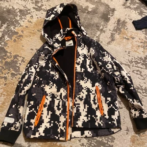 Camouflage vindjacka från Wall Ride - Snygg vindjacka från Wall Ride med svart och vit digitalt camouflagemönster, orange dragkedjor och huva. Jackan är vattenavvisande och har mjukt fleecefoder på insidan. Perfekt för dig som gillar streetstyle och funktionella detaljer. Passar för barn 12 - 13 år. Storlek 158. Jackan är i väldigt bra skick. Pris kan diskuteras! Endast seriösa köpare, tack! ☺️ 