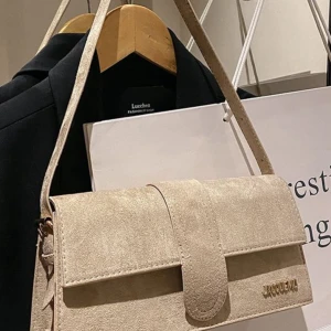 Beige axelväska från Jacquemus - Snygg beige axelväska från Jacquemus i mocka med stilren, rektangulär form och guldfärgad logga på framsidan. Väskan har ett lock med bred rem och justerbar axelrem, perfekt för att bära dina essentials med stil.