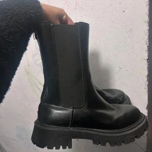 Svarta boots - Säljer ett par svarta chelsea boots med chunky sula och dragkedja bak. Skorna har elastiska paneler på sidorna och är tillverkade i syntetmaterial med matt finish. Perfekta för dig som gillar en edgy och trendig stil.