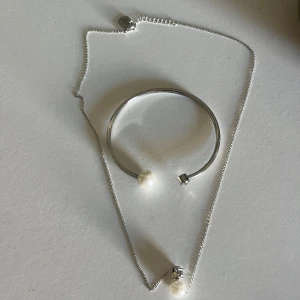 Silverfärgat armband och halsband med pärlor - Edblad - Säljer ett stilrent set med ett silverfärgat armband och matchande halsband. Båda smyckena har en vit pärldetalj som ger en elegant touch. Armbandet är öppet och minimalistiskt medan halsbandet har en tunn kedja och en hängande pärla.