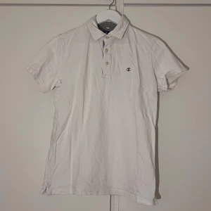 Vit pikétröja från Champion - Vit pikétröja från Champion i klassisk modell krage, knappar fram och ett grått C på framsidan.