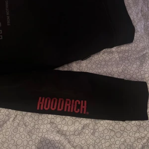 Svart hoodie från Hoodrich med tryck - Svart hoodie från Hoodrich i storlek M med rött och svart tryck på bröstet, ryggen och ena ärmen. Tröjan har huva med snörning och en stor magficka. Perfekt för dig som gillar streetwear och vill sticka ut med en snygg logga.