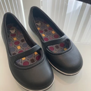 Svarta Crocs ballerinaskor med rem - Svarta ballerinaskor från Crocs med en klassisk rund tå och en rem över foten. Skorna är gjorda i syntetmaterial och har en färgglad innersula med geometriskt mönster i lila, vinrött och gult. Perfekta för dig som vill ha bekväma och stilrena skor.