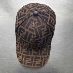 Fendi keps med FF-mönster - Snygg keps från Fendi med det klassiska FF-logomönstret i svart och brunt. Kepsen har böjd skärm, justerbar rem bak och är tillverkad i bomull. Insidan har Fendi-logga på banden och etikett. Perfekt för dig som gillar lyxiga streetwear-vibes.