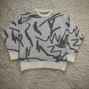 Beige och grå stickad tröja med mönster - Säljer en oversized stickad tröja i beige med grått abstrakt mönster. Tröjan har rund halsringning, ribbade muddar och är långärmad. Perfekt för dig som gillar en chill och trendig look.