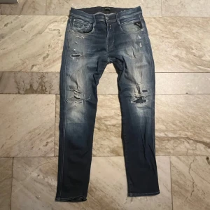 Replay blå slitna jeans slim fit - Säljer ett par blå jeans från Replay med slim fit och slitna detaljer framtill. Jeansen har klassisk femficksdesign, läderpatch bak och snyggt tvättad finish. Perfekta för dig som gillar en avslappnad stil.