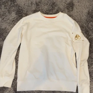 Vit sweatshirt från Moose Knuckles - Säljer en clean vit sweatshirt från Moose Knuckles med guldiga metalldetaljer på ärmen och diskret logga. Tröjan har rund halsringning, långa ärmar och är tillverkad i mjuk bomull. Perfekt för dig som gillar stilrena och exklusiva plagg.