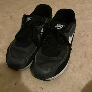 Nike Air Max svarta och grå sneakers - Säljer ett par Nike Air Max sneakers i svart och grått med vit sula och klassisk vit swoosh på sidan. Skorna har snörning och en sportig, chunky design. Ovandelen är i syntet och textil, perfekt för dig som gillar streetwear och bekväma skor.