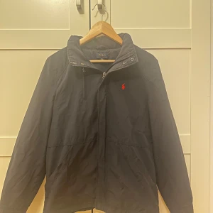 Svart vindjacka Polo Ralph Lauren - Svart vindjacka från Polo Ralph Lauren i storlek XL men passar 175-185cm. Klassisk design med dragkedja framtill, hög krage och röd broderad logga på bröstet. Jackan har långa ärmar och är tillverkad i ett lätt och smidigt material, perfekt för blåsiga dagar.