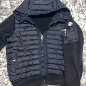 Svart Moncler cardigan med huva - Svart cardigan från Moncler med quiltad framsida, huva och ärmficka med dragkedja. Jackan har Moncler-logga på ärmen och kombinerar dunfylld front med stickade ärmar och rygg. Perfekt för dig som vill ha en sportig och stilren look. Passar M också