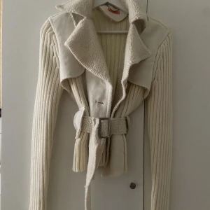 Beige teddyjacka med bälte och stickade ärmar - Snygg beige teddyjacka med bred krage i fuskpäls, stickade ribbade ärmar och kropp, samt detaljer i mockaimitation. Jackan har bälte i midjan och knappar framtill för en cool och mysig look. Perfekt för dig som gillar lager på lager och vill ha något unikt. Passar xs-s