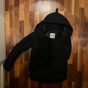 Svart pufferjacka från Kenzo - Svart pufferjacka från Kenzo med huva och diskret logga på ärmen. Jackan har dragkedja och knappar framtill, samt fodrad insida med Kenzo-tryck. Perfekt för kalla dagar och har en clean, modern look.