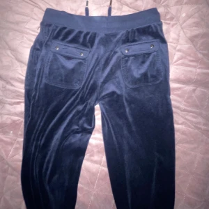 Mörkblå mjukisbyxor från True Couture - Säljer ett par mörkblå mjukisbyxor från True Couture med broderad logga på ena benet. Byxorna har snörning i midjan, stora fickor fram och bak med knappdetaljer. Materialet är mjukt och glansigt, perfekt för chill dagar.