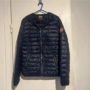 Mörkblå dunjacka från Canada Goose - Snygg mörkblå dunjacka från Canada Goose med horisontella sömmar, huva och dragkedja framtill. Jackan har två fickor med dragkedja och klassisk logotyp på ärmen. Perfekt för kyliga dagar och enkel att matcha med olika outfits.