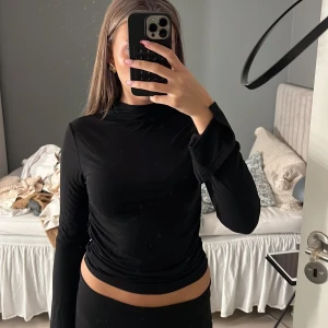 Svart långärmad polotopp med rynk - Säljer en svart långärmad polotopp med croppad passform och snygga rynkade detaljer i sidorna. Toppens material är stretchigt och mjukt, perfekt för en stilren look. Hög hals och långa ärmar med lätt utsvängda avslut.