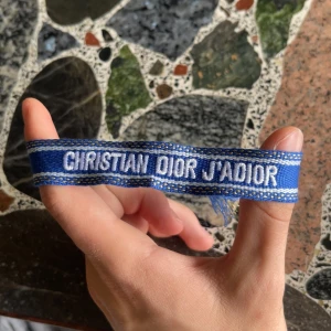 Blå Christian Dior J'Adior armband - Snyggt blått armband från Christian Dior med texten 'J'Adior' broderad i vitt. Armbandet har dekorativa tofsar i ändarna och detaljer i guld. Perfekt accessoar för att lyfta din outfit med lite lyxig vibe.