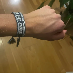 Christian Dior blått tygarmband - Snyggt blått armband  med vit text och detaljer. Armbandet är gjort i tyg och har flätade snören med tofsar i ändarna. Perfekt accessoar för att lyfta din outfit med en exklusiv touch och cool design.