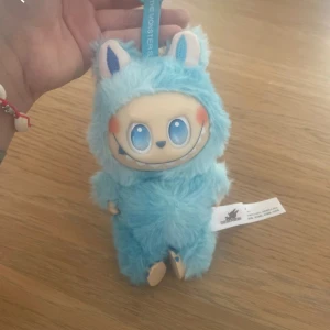 Pop Mart The Monsters Blue Plush - Söt Pop Mart The Monsters mjukisfigur i blått fluffigt tyg med stora blå ögon och ett glatt leende. Figuren är i mycket fint skick utan synligt slitage, perfekt för samlare eller som present. Originaletiketter sitter kvar.