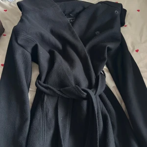 Svart omlottkappa med bälte från MISU - Medellång Svart kappa med luva från Amisu med knappar innanför. Använd ett par gånger så den har hunnit bli lite nopprig men ingen som syns mycket, Storlek 40 men passar också jätte fint för storlek M och S
