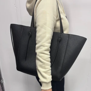 Svart shoppingväska från GinaTricot - Stor svart shoppingväska med stilren design och långa handtag. Väskan är tillverkad i strukturerat skinn och har minimalistiska detaljer med knyt på sidorna. Rymlig insida med magnetknapp för stängning, perfekt för dig som gillar att bära med dig mycket.