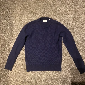 Mörkblå stickad tröja från Bläck - Säljer en mörkblå stickad tröja från Bläck i storlek M men sitter som XS.Tröjan har ett snyggt diskret mönster framtill och är ribbstickad vid hals, ärmslut och nederkant. Perfekt för kyliga dagar och enkel att matcha med jeans eller chinos.
