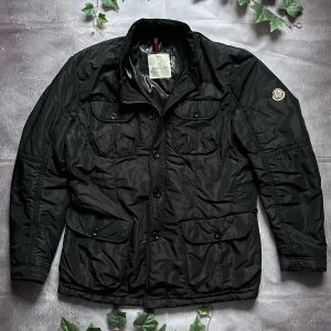 Moncler Austin Jacket – Svart storlek 4 (Äkta) - 🎨 Färg: Svart 📏 Storlek: 4 (motsvarar L) 📐 Mått: Axel till axel ca 47 cm, längd ca 70 cm, ärmlängd ca 65 cm ♻️ Skick: Väldigt bra (8,5–9/10) – välvårdad med minimala tecken på användning 🧵 Material: Nylon (yttertyg), lätt fyllning, logodetalj på ärm  ‼️Skickas dagen efter köpet genomförs‼️