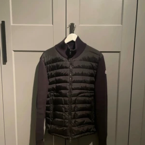 Svart quiltad höstjacka  från Moncler - Snygg svart pufferjacka från Moncler med quiltad front och ärmar i stickad kvalitet. Jackan har hög krage, dragkedja framtill och Moncler-logga på ärmen. Perfekt för dig som vill ha en stilren och exklusiv look.