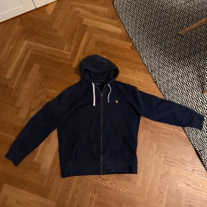 Marinblå hoodie från Polo Ralph Lauren - Marinblå hoodie från Polo Ralph Lauren med dragkedja, huva med vita snören och klassisk gul logga på bröstet. Tillverkad i mjuk bomullsblandning med fickor framtill. Perfekt för en chill och stilren look.